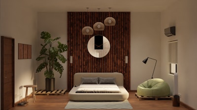 A modern living room features a low-profile sofa bed in the center, set against a wooden panel wall with a round decorative mirror. Above the sofa hang several pendant lights. To the left, a large potted plant adds a touch of greenery, next to a small wooden stool. On the right, a stylish floor lamp stands beside a green, plush armchair. The room also has an air conditioning unit and a window with a rolled-up blind.