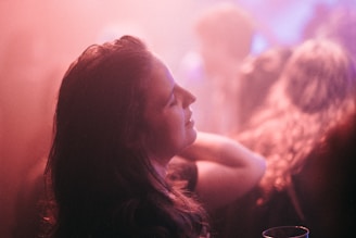 A person with closed eyes appears lost in thought or immersed in the atmosphere of what seems to be a party or club environment. The background is hazy with a pink and purple hue, creating a dreamy and serene ambiance. The person has their head slightly tilted back, with surrounding out-of-focus figures suggesting a lively setting.