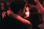 A couple embraces closely in a dimly lit setting, sharing an intimate and joyful moment. The focus is on their faces, illuminated by warm, soft lighting, suggesting an atmosphere of affection and connection.