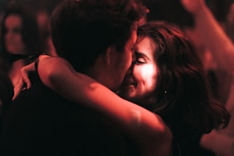 A couple embraces closely in a dimly lit setting, sharing an intimate and joyful moment. The focus is on their faces, illuminated by warm, soft lighting, suggesting an atmosphere of affection and connection.