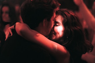A couple embraces closely in a dimly lit setting, sharing an intimate and joyful moment. The focus is on their faces, illuminated by warm, soft lighting, suggesting an atmosphere of affection and connection.