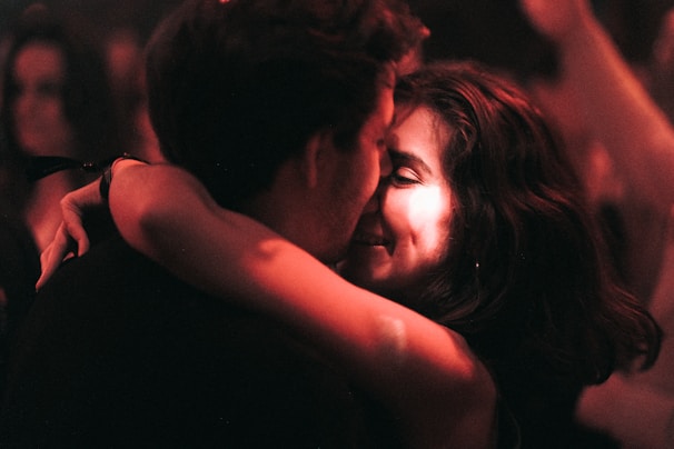 A couple embraces closely in a dimly lit setting, sharing an intimate and joyful moment. The focus is on their faces, illuminated by warm, soft lighting, suggesting an atmosphere of affection and connection.