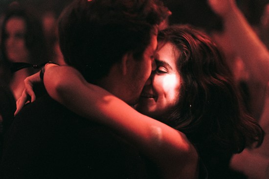 A couple embraces closely in a dimly lit setting, sharing an intimate and joyful moment. The focus is on their faces, illuminated by warm, soft lighting, suggesting an atmosphere of affection and connection.