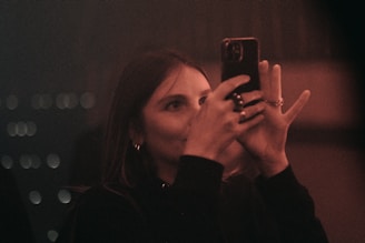 A woman is holding a smartphone in her hands. The lighting is dim, giving the scene a warm, moody atmosphere. Her face is partially illuminated by the phone's screen, and she appears focused on capturing a photograph.