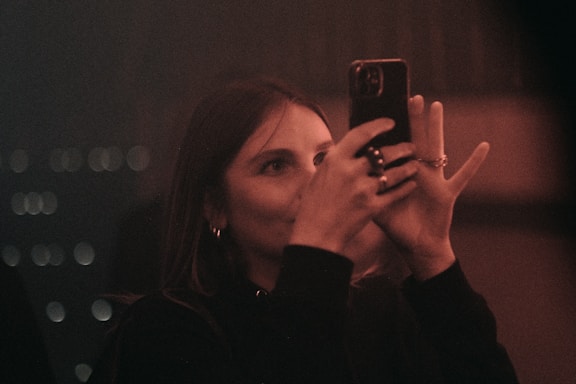 A woman is holding a smartphone in her hands. The lighting is dim, giving the scene a warm, moody atmosphere. Her face is partially illuminated by the phone's screen, and she appears focused on capturing a photograph.