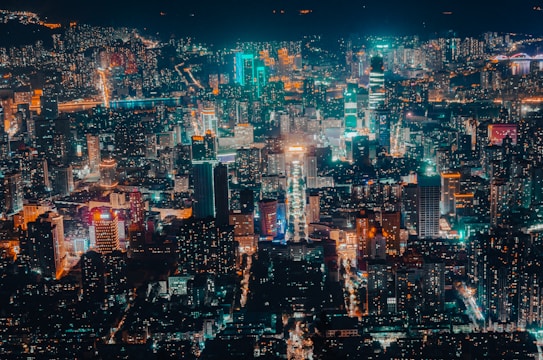 A bustling urban cityscape at night with tall buildings and glowing lights in a spectrum of colors. Streets are visible with bright neon illumination, and the overall scene conveys a sense of vibrancy and energy.