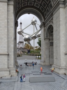 A view through an ornate stone archway reveals a miniature scene, featuring iconic structures like the Eiffel Tower and the Atomium. Small figures of people are scattered across the paved plaza, creating a bustling atmosphere.