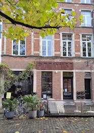 A brick building with large rectangular windows and a sign above the entrance displaying the word 'OPTIMIST'. In front of the building, there is a collection of potted plants, two parked bicycles, and a metal bicycle rack. The scene is shaded by green and yellow leaves from a tree overhead, and a cobblestone pavement runs along the front.