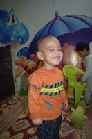 A child wearing an orange sweatshirt is smiling broadly, standing in a play area decorated with colorful murals and a patterned carpet. Another child is visible in the background interacting with a green chair. A plush caterpillar toy lies on the carpet.