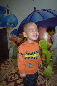 A child wearing an orange sweatshirt is smiling broadly, standing in a play area decorated with colorful murals and a patterned carpet. Another child is visible in the background interacting with a green chair. A plush caterpillar toy lies on the carpet.