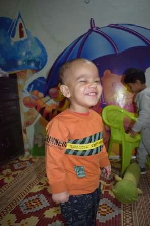 A child wearing an orange sweatshirt is smiling broadly, standing in a play area decorated with colorful murals and a patterned carpet. Another child is visible in the background interacting with a green chair. A plush caterpillar toy lies on the carpet.