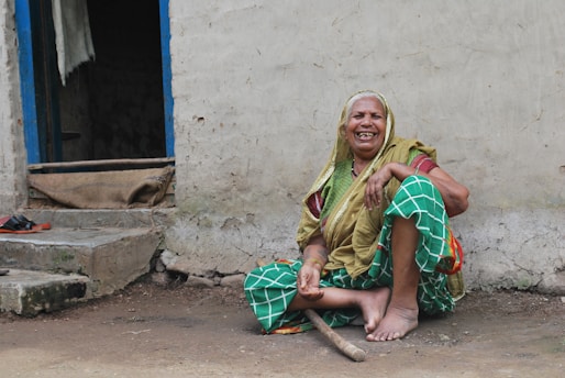 An elderly woman with a cheerful expression sits on the ground near the entrance of a weathered building. She wears a colorful sari and headscarf, with hues of green, yellow, and red. The doorframe is painted blue, and a pair of shoes and a burlap sack are visible on the steps leading inside. The building’s exterior shows signs of wear and age.