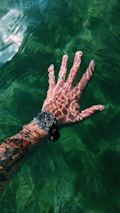 A tattooed hand submerged partially in clear green water, with sunlight creating intricate patterns on the skin. The hand is wearing a wristwatch and a silver ring.