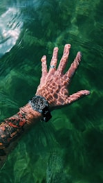 A tattooed hand submerged partially in clear green water, with sunlight creating intricate patterns on the skin. The hand is wearing a wristwatch and a silver ring.