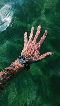 A tattooed hand submerged partially in clear green water, with sunlight creating intricate patterns on the skin. The hand is wearing a wristwatch and a silver ring.