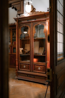 An intricately carved wooden cabinet is displayed with glass doors, housing various antique books and manuscripts. The cabinet is set against a warmly lit room with wooden floors and classical decor. A large, vintage light fixture hangs above the cabinet, casting a soft glow on the woodwork.