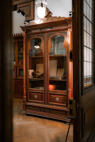 An intricately carved wooden cabinet is displayed with glass doors, housing various antique books and manuscripts. The cabinet is set against a warmly lit room with wooden floors and classical decor. A large, vintage light fixture hangs above the cabinet, casting a soft glow on the woodwork.