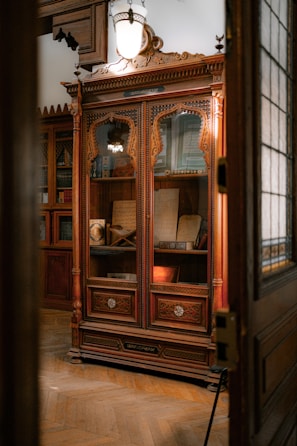 An intricately carved wooden cabinet is displayed with glass doors, housing various antique books and manuscripts. The cabinet is set against a warmly lit room with wooden floors and classical decor. A large, vintage light fixture hangs above the cabinet, casting a soft glow on the woodwork.
