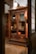 An intricately carved wooden cabinet is displayed with glass doors, housing various antique books and manuscripts. The cabinet is set against a warmly lit room with wooden floors and classical decor. A large, vintage light fixture hangs above the cabinet, casting a soft glow on the woodwork.