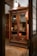 An intricately carved wooden cabinet is displayed with glass doors, housing various antique books and manuscripts. The cabinet is set against a warmly lit room with wooden floors and classical decor. A large, vintage light fixture hangs above the cabinet, casting a soft glow on the woodwork.