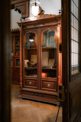 An intricately carved wooden cabinet is displayed with glass doors, housing various antique books and manuscripts. The cabinet is set against a warmly lit room with wooden floors and classical decor. A large, vintage light fixture hangs above the cabinet, casting a soft glow on the woodwork.