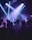 A crowded nightclub scene with people dancing and raising their hands in the air. Purple and blue stage lights illuminate the crowd, creating a lively and energetic atmosphere. Many individuals are wearing casual clothing, and some are taking photos or videos with their phones.
