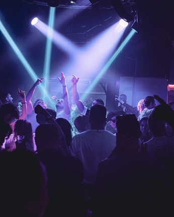 A crowded nightclub scene with people dancing and raising their hands in the air. Purple and blue stage lights illuminate the crowd, creating a lively and energetic atmosphere. Many individuals are wearing casual clothing, and some are taking photos or videos with their phones.