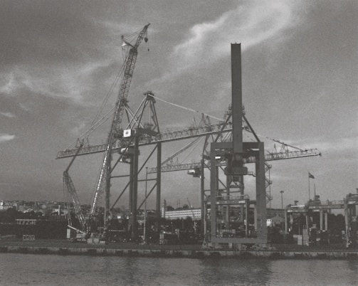 Cranes tower over a shipyard or dock area, with tall structures silhouetted against a cloudy sky. The scene is industrial, featuring large machinery and equipment used for loading and unloading ships. The waterfront is bordered by a quay where various smaller structures and a flagpole can be seen.