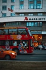 A red double-decker bus with artwork and advertisements travels down the street with a modern urban building in the background. The Marriott hotel logo is visible on the building. An orange car is visible in the foreground, driving alongside the bus.