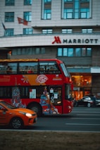 A red double-decker bus with artwork and advertisements travels down the street with a modern urban building in the background. The Marriott hotel logo is visible on the building. An orange car is visible in the foreground, driving alongside the bus.