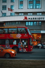 A red double-decker bus with artwork and advertisements travels down the street with a modern urban building in the background. The Marriott hotel logo is visible on the building. An orange car is visible in the foreground, driving alongside the bus.