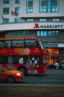 A red double-decker bus with artwork and advertisements travels down the street with a modern urban building in the background. The Marriott hotel logo is visible on the building. An orange car is visible in the foreground, driving alongside the bus.