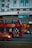 A red double-decker bus with artwork and advertisements travels down the street with a modern urban building in the background. The Marriott hotel logo is visible on the building. An orange car is visible in the foreground, driving alongside the bus.