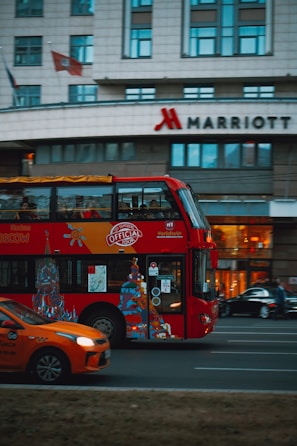 A red double-decker bus with artwork and advertisements travels down the street with a modern urban building in the background. The Marriott hotel logo is visible on the building. An orange car is visible in the foreground, driving alongside the bus.