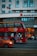 A red double-decker bus with artwork and advertisements travels down the street with a modern urban building in the background. The Marriott hotel logo is visible on the building. An orange car is visible in the foreground, driving alongside the bus.
