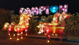 Three illuminated horse sculptures stand in front of a vibrant display with large, colorful letters. The horses are intricately designed with bright red bodies and golden manes, glowing against a nighttime backdrop. The letters spell out a message with a heart symbol, adding to the festive atmosphere.
