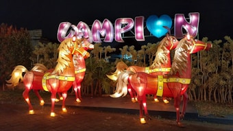Three illuminated horse sculptures stand in front of a vibrant display with large, colorful letters. The horses are intricately designed with bright red bodies and golden manes, glowing against a nighttime backdrop. The letters spell out a message with a heart symbol, adding to the festive atmosphere.