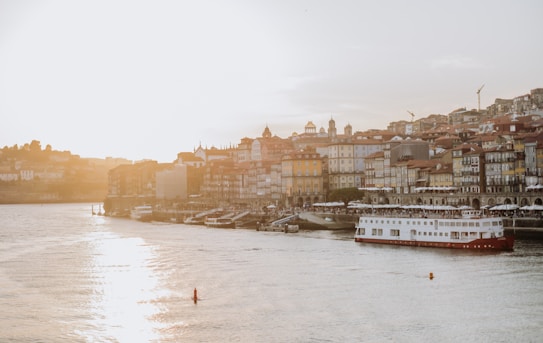 A serene waterfront scene depicts a sunny view of an old town with a row of colorful, historic buildings lining the pier. A large white and red boat is docked along the river, reflecting in the calm water. The sun is prominently setting in the background, casting a warm glow over the entire landscape.