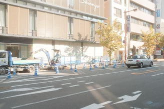 A street scene with road construction taking place. There is a small excavator and a truck parked adjacent to the construction area, which is surrounded by blue and white traffic cones. Several workers are present, wearing safety helmets and vests. The background features a modern building with large windows and wooden paneling. Trees with yellow-brown leaves line the street, indicating an autumn setting. There are also parked and moving cars on the road, with traffic markings clearly visible.