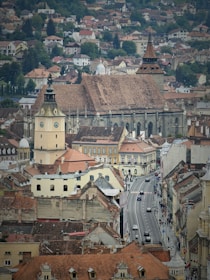 A historic cityscape featuring a large Gothic-style church with a tall steeple, surrounded by a variety of older buildings with red-tiled roofs. The foreground shows a lively street with cars and pedestrians, flanked by classic architecture. The backdrop includes a blend of trees and smaller homes spread across the hilly terrain.