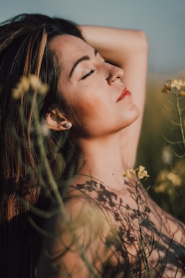 A serene woman with long hair and closed eyes appears relaxed amidst a field of yellow flowers, enjoying sunlight on her face.
