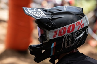 A person is wearing a black motocross helmet adorned with a prominent 100% brand logo in bold red lettering on the strap. The helmet features a visor, protective goggles, and graphic designs in white and subtle colored accents.