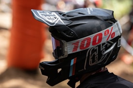 A person is wearing a black motocross helmet adorned with a prominent 100% brand logo in bold red lettering on the strap. The helmet features a visor, protective goggles, and graphic designs in white and subtle colored accents.