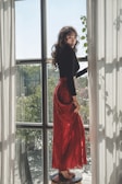 A woman with long curly hair stands on a small stool by a large window. She is wearing a long red skirt and a black top, with natural light streaming through sheer curtains. The view outside shows greenery, adding a serene, nature-inspired backdrop.