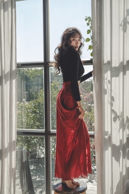 A woman with long curly hair stands on a small stool by a large window. She is wearing a long red skirt and a black top, with natural light streaming through sheer curtains. The view outside shows greenery, adding a serene, nature-inspired backdrop.