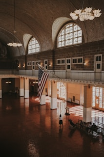 An expansive indoor hall with large arched windows allowing natural light to pour into the space. The room features a high vaulted ceiling adorned with elegant chandeliers. A prominent American flag hangs from the ceiling. The polished, reflective floor adds to the majestic ambiance. A few people are scattered around; one person is pushing a stroller and others are sitting on benches.