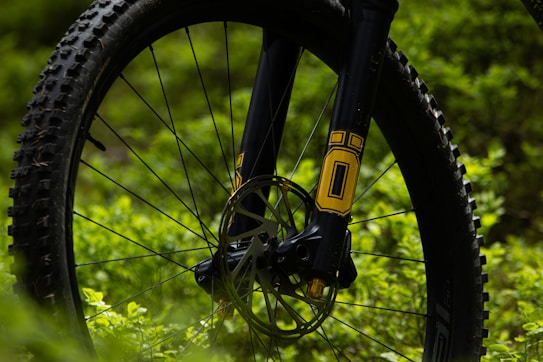A close-up view of a mountain bike tire and suspension fork amidst greenery. The bike components feature black and yellow accents, highlighting the disc brake and tread pattern against the lush background.