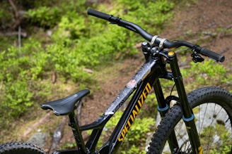 A close-up view of a black mountain bike featuring large tires and a prominent handlebar set against a backdrop of lush green foliage. The bike frame displays the brand name in bold yellow lettering.