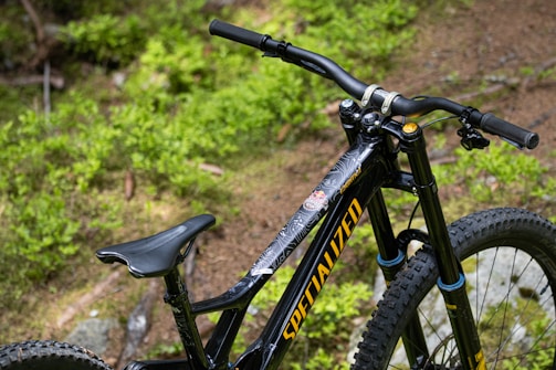 A close-up view of a black mountain bike featuring large tires and a prominent handlebar set against a backdrop of lush green foliage. The bike frame displays the brand name in bold yellow lettering.