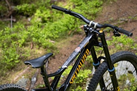 A close-up view of a black mountain bike featuring large tires and a prominent handlebar set against a backdrop of lush green foliage. The bike frame displays the brand name in bold yellow lettering.